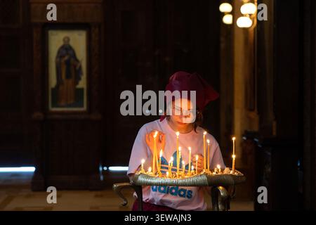 Eine Person mit lila Hut und weißem Hemd hält ein Tablett mit beleuchteten Kerzen in einem schwach beleuchteten Raum mit Holzwänden und religiösen Ikonen. Stockfoto
