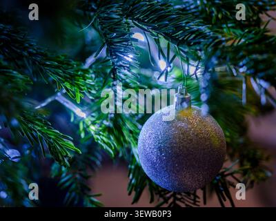 Nahaufnahme einer glitzernden Silberkugel, die an einem Weihnachtsbaum hängt, mit leuchtenden Lichtern und weicher festlicher Atmosphäre Stockfoto