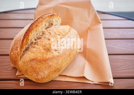 Frisches handwerkliches rustikales Brotbrot mit goldener Kruste auf einer braunen Papiertüte über einem Holztisch, auf dem natürlich gesundes lokales Backen präsentiert wird. Stockfoto