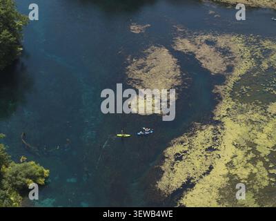 Blick aus der Vogelperspektive auf die natürliche Quelle Blue Eye mit Personen auf Paddleboards, die das klare blaue Wasser inmitten von aquatischer Vegetation in Finiq, Vlor, navigieren Stockfoto