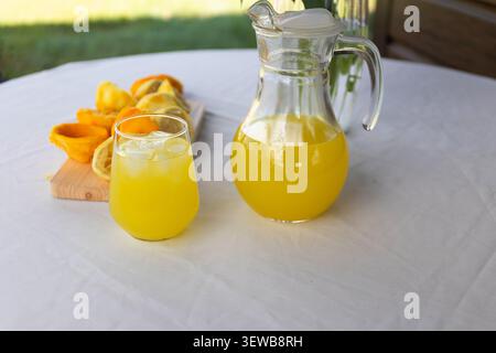 Krug und Becher aus klarem Glas gefüllt mit leuchtend orangener Limonade auf weißer Tischdecke neben halbierten Zitrusstücken, die frische Sommererfrischung zeigen Stockfoto
