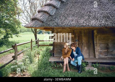 Ein entspanntes Paar in Sommeroutfits sitzt zusammen auf einer Bank in der Nähe eines charmanten Cottages Stockfoto