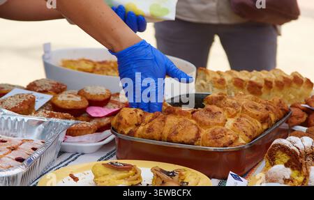 Individual mit blauen Handschuhen wählt Backwaren von einem Marktstand aus, gefüllt mit verschiedenen Backwaren, Brot und Desserts auf Tischen Stockfoto