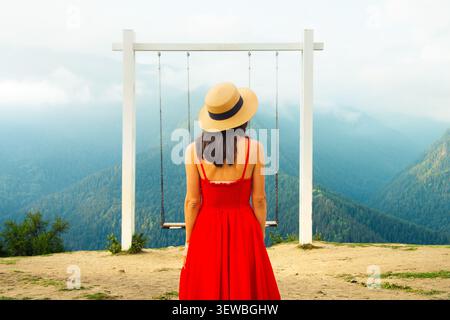 Elegante Frau in fließendem rotem Kleid, die zwischen Bergschaukeln mit Blick auf das nebelige Waldtal in grünen Bergen steht. Konzeptfreiheit Reise uns Stockfoto