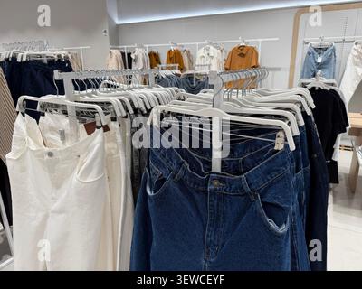 Verschiedene Damenbekleidung, darunter Denim-Jeans und weiße Hosen an Kleiderbügeln, in einem modernen Ladengeschäft Stockfoto