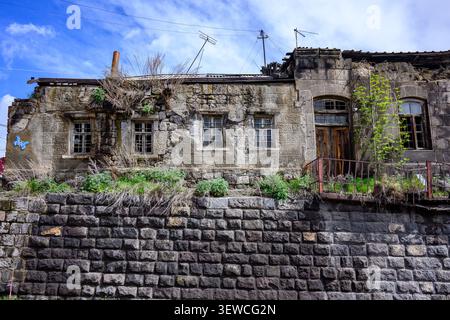Altes Steinhaus mit Moos und wilden Pflanzen. Verwitterte Wände, kaputte Fenster, bewachsene Vegetation. Steinfangmulde vorne. Blauer Himmel mit Wolken Stockfoto