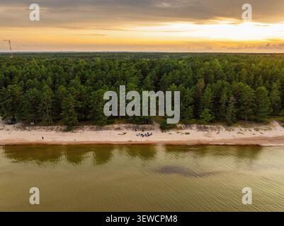Luftaufnahme einer Gruppe von Menschen, die auf dem Sand sitzen und während der goldenen Stunde in Melnsils ein Akustikgitarrenkonzert in der Nähe eines dichten Kiefernwaldes hören, Stockfoto