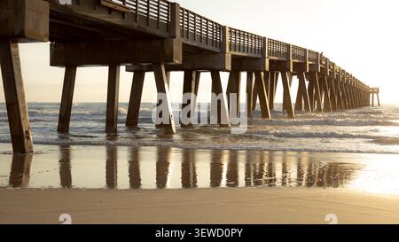 Wunderschönes goldenes Sonnenaufgangslicht, das auf den feuchten Sand- und Betonsäulen des Angelpiers Jacksonville Beach reflektiert. Stockfoto