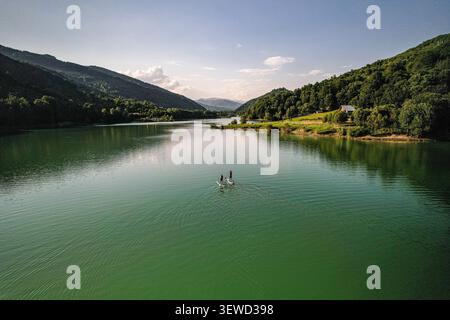 Aus der Vogelperspektive von zwei Personen auf Stehpaddelbrettern auf einem ruhigen See umgeben von üppig grünen Bergen in Tesila, Prahova, Rumänien. Stockfoto