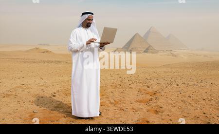 Lächelnder arabischer Geschäftsmann in weißer Kandura mit Laptop in der ägyptischen Wüste in der Nähe der Pyramiden von Gizeh, die Remote Work Connectivity und Middle darstellen Stockfoto