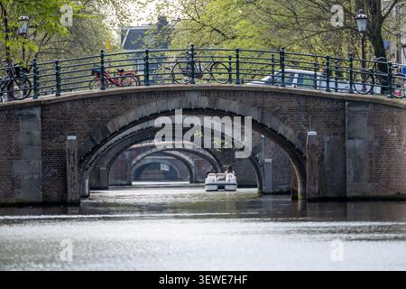 Backsteinkanalbrücke mit mehreren Bögen und Fahrrädern darüber, die ein kleines Tretboot auf einem Amsterdamer Kanal in Nordholland, Niederlande, umrahmt. Stockfoto