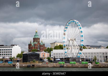 Helsinki SkyWheel und die rote Backstein-Uspenski-Kathedrale unter einem dramatischen, bewölkten Himmel, gesehen vom Hafen in Finnland. Stockfoto