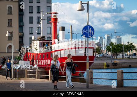 Zwei junge Frauen schlendern entlang einer sonnigen Hafenpromenade in Helsinki, mit einer markanten roten Bootsbar im Hintergrund. Stockfoto