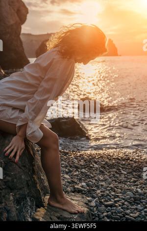 Frau Sonnenuntergang Strand sitzt an einem felsigen Ufer in einem weißen Hemd und genießt die goldene Stunde über dem Meer Stockfoto