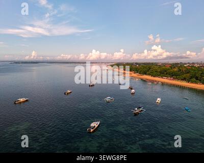 Blick aus der Vogelperspektive auf Sanur Beach mit traditionellen Auslegerbooten, die auf ruhigem türkisfarbenem Wasser entlang der Sandküste in Kecamatan Denpasar Selatan schwimmen, Stockfoto