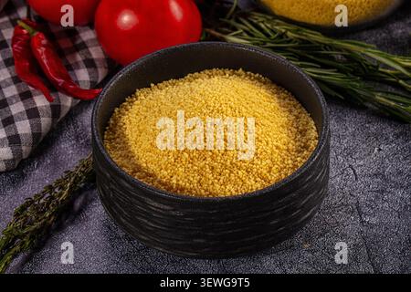 Nahaufnahme von gelbem Couscous in schwarzer Keramikschale, umgeben von roten Chilischoten, Tomaten, Rosmarin, Thymian auf kariertem Tuch. Dunkel strukturierter Hintergru Stockfoto