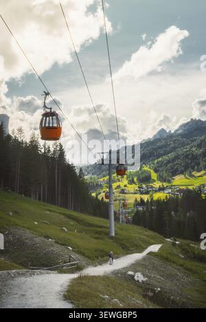 Dolomiten, Italien - 27. juli 2026: Gondelbahn Colfosco Orange über saftig grüne Almwiesen Wanderweg Tal Stadt Kiefernwald towe Stockfoto