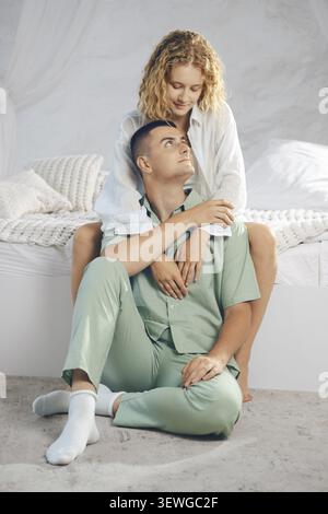 Mann und Frau teilen einen zärtlichen Moment im Schlafzimmer. Sie sitzen auf dem Boden mit Bett und Decken im Hintergrund. Sonnenlicht füllt den Raum. Das Paar genießt eine ruhige Zeit Stockfoto