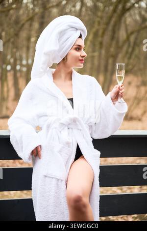Junge Frau, die in einen weichen Mantel mit Handtuch-Turban gewickelt ist und Sekt auf einer Bank im Wald hält. Konzept von Wellness-Rückzugsort, Selbstpflege, Entspannung und Luxus-Zuflucht Stockfoto