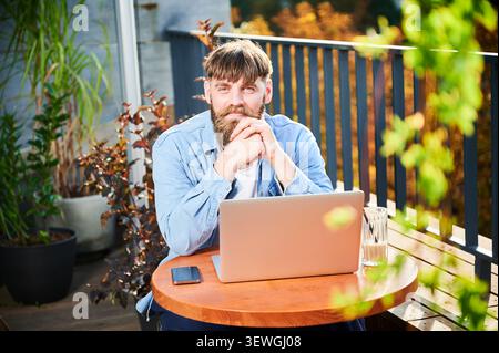 Porträt eines Mannes mit Bart und Laptop im Sitzbereich im Freien, umgeben von Laub, warmem Licht, Smartphone und städtischer Terrassenatmosphäre, geeignet für Fernarbeit Stockfoto
