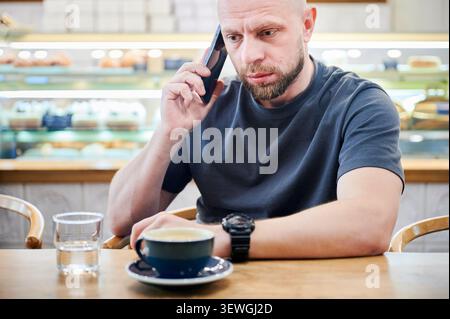 Mann, der am Café-Tisch mit einer Tasse Kaffee, ernsthaftem Ausdruck, Kommunikationskonzept, Geschäftsanruf, Fernarbeit und modernem Lebensstil in Innenräumen spricht Stockfoto