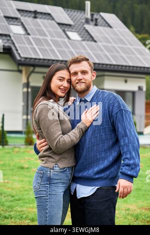 Ein Paar, das sich in der Nähe des Hauses mit Solarpaneelen umschließt und in die Kamera lächelt. Glückliche Hausbesitzer, die erneuerbare Energien, umweltfreundliches Zuhause und nachhaltiges Lifestyle-Konzept nutzen Stockfoto