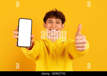 Ein lächelnder Kerl hält einen leeren Smartphone-Bildschirm und gibt die Daumen hoch Stockfoto
