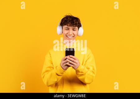 Lächelnder junger Mann in gelbem Hoodie mit weißen Kopfhörern, während er das Smartphone benutzt Stockfoto