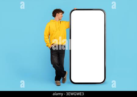 Entspannter junger Mann in gelbem Hoodie, der sich auf ein großes leeres Smartphone-Modell stützt Stockfoto
