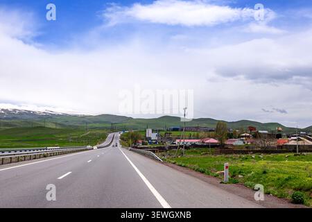 Eine lange, gerade Straße zieht sich durch die grüne Landschaft unter blauem Himmel mit weißen Wolken. Hügel mit grünem Gras und Bäumen säumen den Horizont Stockfoto