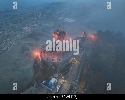 Blick aus der Vogelperspektive auf den Guaita-Turm, die historische Festung auf dem Titano, umgeben von Nebel und beleuchtet von warmen Lichtern am Abend in Citta Stockfoto