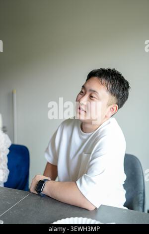 Junger Mann sitzt entspannt und nachdenklich am Tisch. Er blickt sanft, die Augen geschlossen, in ruhiger Besinnung. Schlichtes weißes T-Shirt, lässig, dezent. Dunkle Uhr Stockfoto