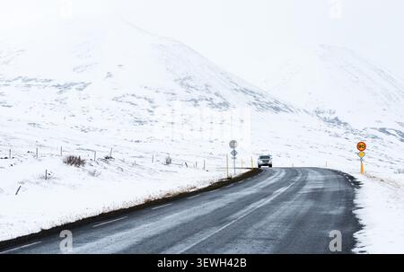 Weißer geländewagen, der auf einer Asphaltstraße durch eine riesige verschneite Landschaft mit weit entfernten Bergen im Winter fährt. Schneesturm, island Stockfoto