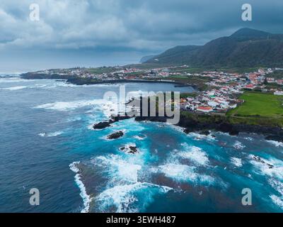 Blick aus der Vogelperspektive auf den Strand von Mosteiros mit dunklen vulkanischen Felsen, stürzenden türkisfarbenen Wellen und die Küstenstadt eingebettet in grüne Hügel in Mosteiros, ACOR Stockfoto