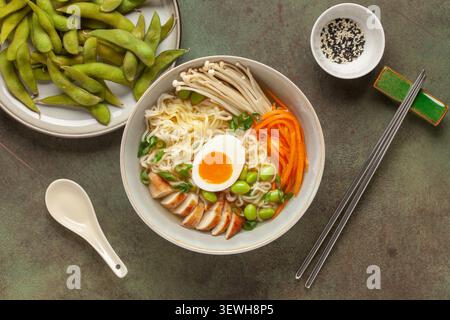 Asiatische Ramen-Nudeln mit Hähnchenscheibe, Enoki-Pilzen, Karotten, Edamame-Bohnen und Ei in einer Schüssel. Draufsicht Stockfoto
