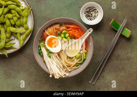 Asiatische Ramen-Nudeln mit Hähnchenscheibe, Enoki-Pilzen, Karotten, Edamame-Bohnen und Ei in einer Schüssel. Draufsicht Stockfoto