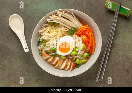 Asiatische Ramen-Nudeln mit Hähnchenscheibe, Enoki-Pilzen, Karotten, Edamame-Bohnen und Ei in einer Schüssel. Draufsicht Stockfoto