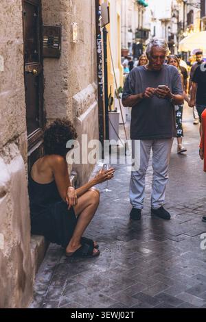 Ein Mann, der sein Smartphone überprüft, und eine Frau, die auf einer engen Altstadtstraße in Ortigia Syrakus auf Sizilien ruht. Italienisches Straßenleben in einem historischen Medit Stockfoto