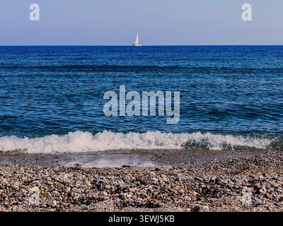 Die Wellen krachen sanft am felsigen Strand, während ein weit entferntes Segelboot an einem klaren Tag in Rethymno, Kreta, über das blaue Meer fährt. Stockfoto