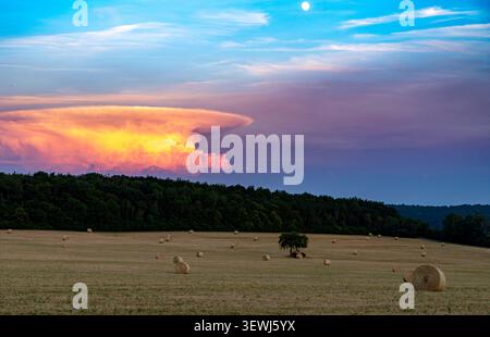 Futuristischer Sonnenuntergang über erntetem Feld mit runden Heuballen, einsamen Bäumen und einer dramatischen Sturmwolke, die einem riesigen Raumschiff ähnelt Stockfoto