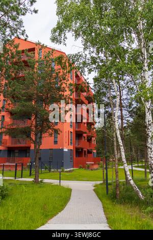 Modernes, helles, rotes mehrstöckiges Apartmentgebäude mit geometrischen Balkonen, das sich neben einem grünen öffentlichen Park und Fußgängerweg befindet. Stockfoto