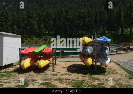 Eine große Aufnahme von mehrfarbigen Plastikkayaks und Kanus, die auf Metallregalen an einer Verleihstation in der Nähe eines malerischen Bergsees gelagert werden. Ein dichter grüner Kiefernfurt Stockfoto