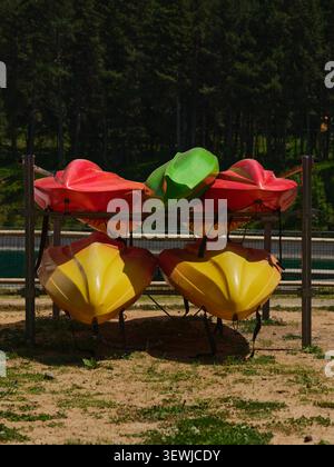 Eine große Aufnahme von mehrfarbigen Plastikkayaks und Kanus, die auf Metallregalen an einer Verleihstation in der Nähe eines malerischen Bergsees gelagert werden. Ein dichter grüner Kiefernfurt Stockfoto
