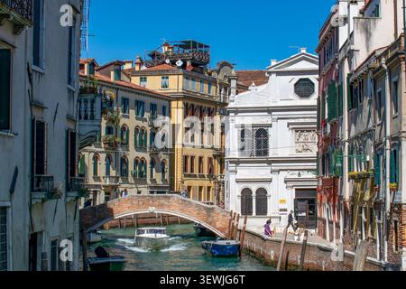 Malerische Kanalszene in Venedig, Italien, mit einer Backsteinbrücke, historischen Gebäuden und einem Motorboot, das unter einem klaren blauen Himmel vorbeifährt. Stockfoto