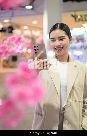 Ein vertikales Nahporträt einer fröhlichen jungen südostasiatischen thailändischen Geschäftsfrau aus der Isan-Region, die mit einem breiten Lächeln auf ihr Handy hinunterblickt Stockfoto