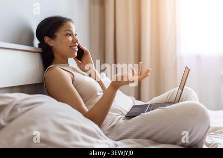 Glückliche schwarze Frau, die auf dem Bett mit Laptop liegt, während sie auf dem Smartphone spricht Stockfoto