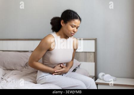 Afroamerikanerin, die auf dem Bett sitzt und den Magen mit Schmerzen hält Stockfoto