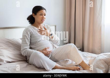 Schwangere schwarze Frau, die auf dem Bett sitzt und Brust und Bauch hält Stockfoto