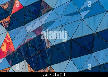 Dynamische architektonische Fassade mit dreieckigen Paneelen, symmetrischem Design, leuchtenden Blautönen, Reflexionen der urbanen Umgebung, Lichtspiel Stockfoto