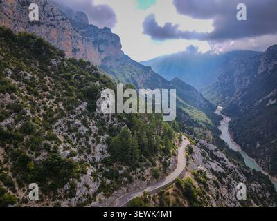 Ein Blick aus der Vogelperspektive zeigt eine gewundene Straße, die sich an einem steilen, felsigen Berghang anschmiegt, der mit grünem Gestrüpp bedeckt ist und durch den der türkisfarbene Verdon River fließt Stockfoto